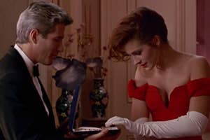 Man in tuxedo presents jewelry box to woman in red off-the-shoulder gown with white gloves, intimate setting