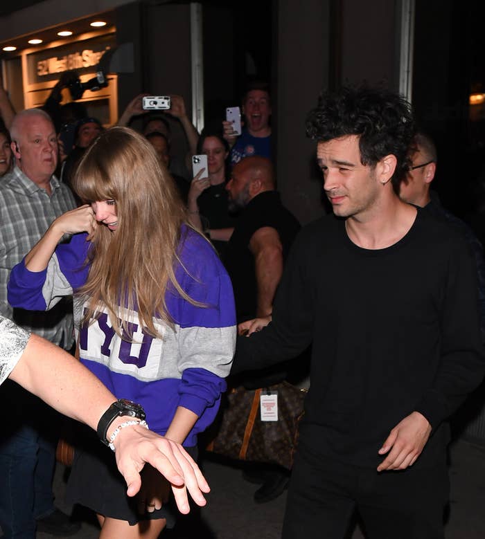 Taylor Swift in a purple sweatshirt and shorts exits a building with Matty Healy in black attire. Fans surround them