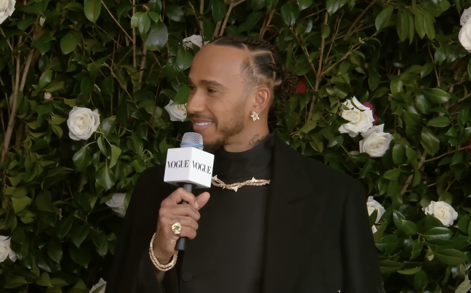 Lewis Hamilton in a black suit with jewelry, holding a mic, interview backdrop
