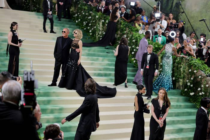 Celebrities in elegant attire on a staircase at a gala, surrounded by photographers and onlookers