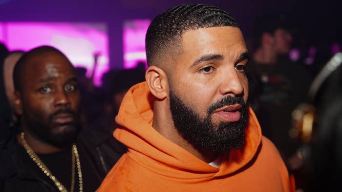 Drake in an orange hoodie at an event, with another man in the background