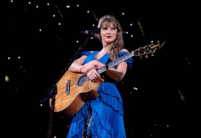 Taylor Swift on stage with a guitar, wearing a sparkly blue dress with ruffles. She&#x27;s smiling under a star-like lit backdrop