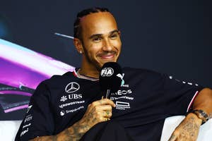Lewis Hamilton seated, smiling, holding a microphone, wearing Mercedes F1 team gear