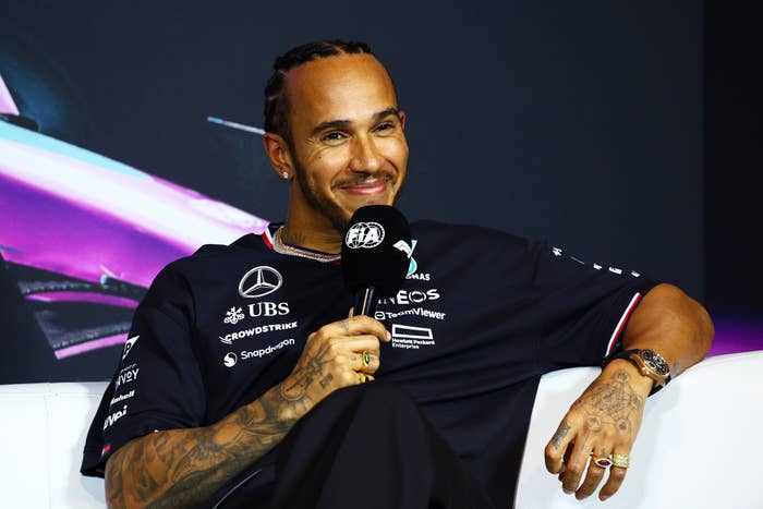 Lewis Hamilton seated, smiling, holding a microphone, wearing Mercedes F1 team gear