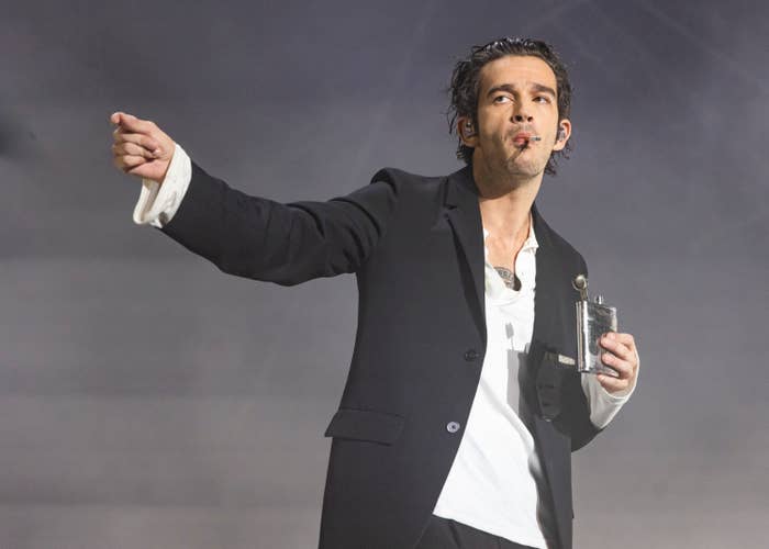 Matty Healy in suit holding a microphone, gesturing with his arm extended, against a smoky background