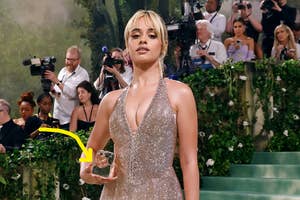 Camila Cabello in a sparkling gown with plunging neckline, holding an ice cube, on the Met Gala stairs. Photographers in background