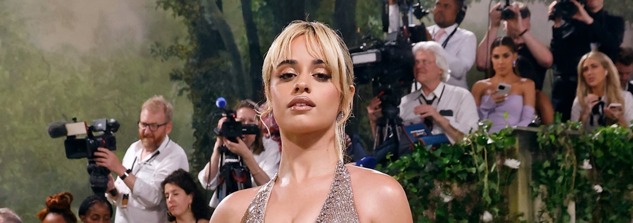 Camila Cabello in a sparkling gown with plunging neckline, holding an ice cube, on the Met Gala stairs. Photographers in background