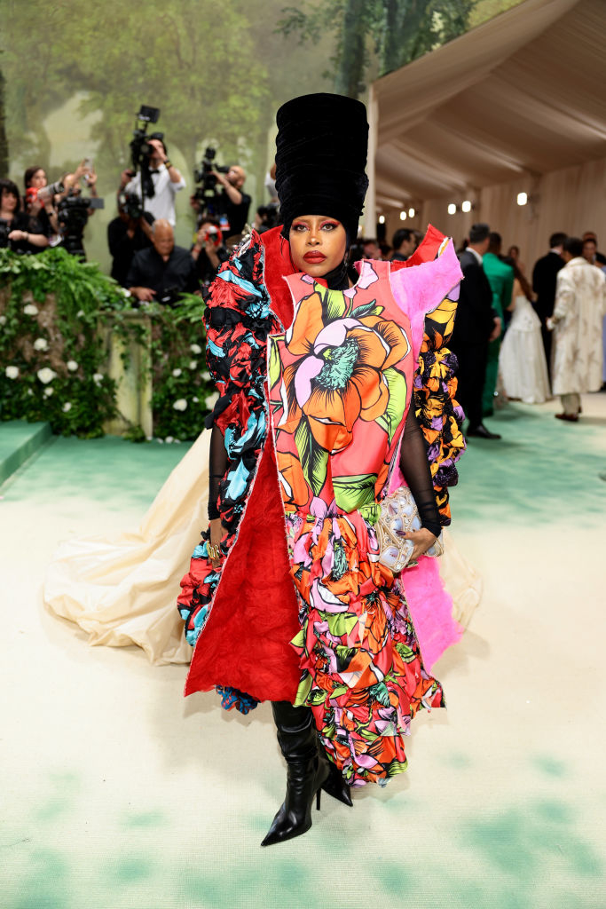 Erykah Badu at an event in a multi-textured floral outfit with a tall black headpiece, photographers in the background