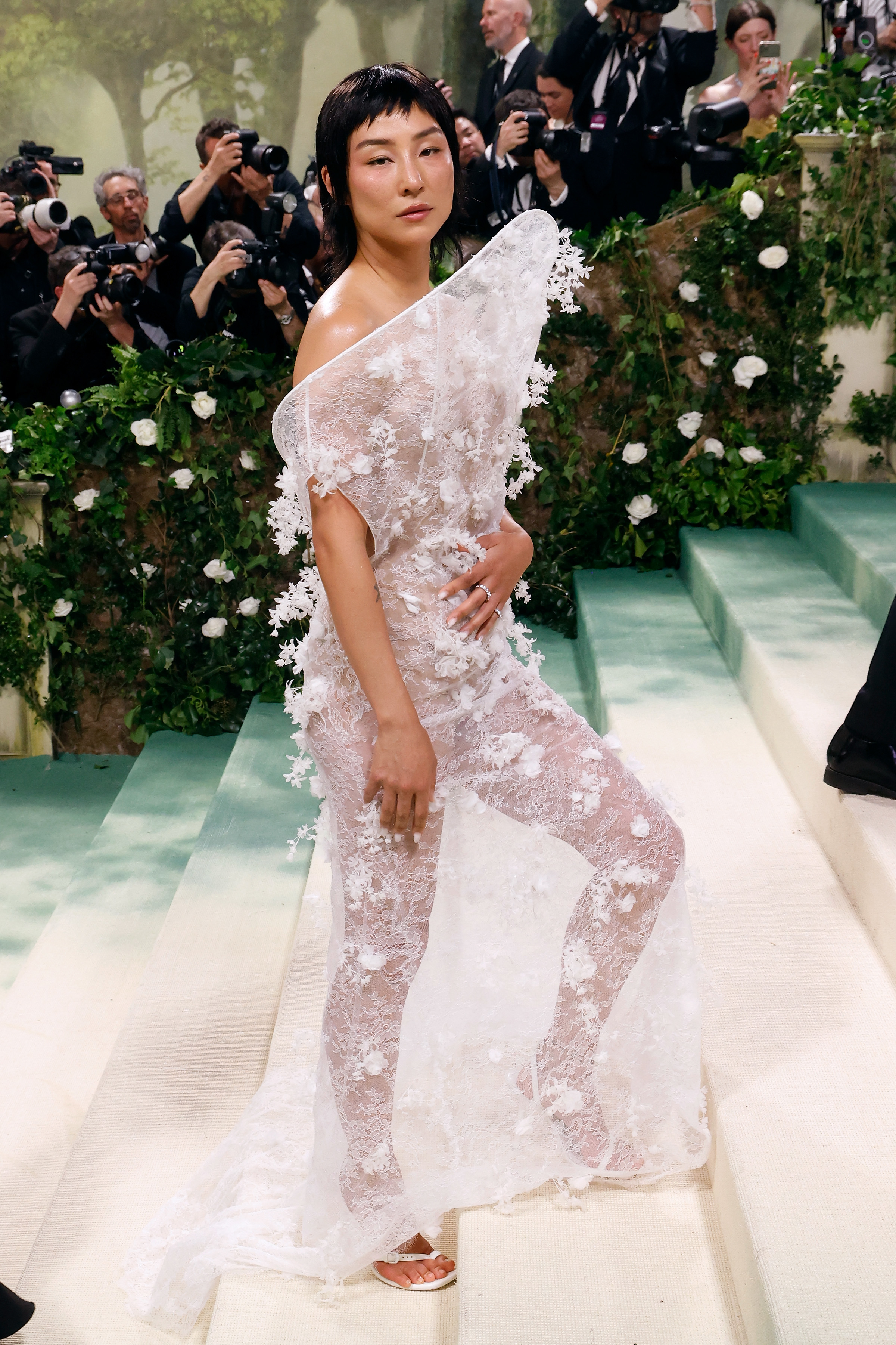 Greta Lee standing on the steps of the Met at the 2024 Met Gala wearing a sheer, floral outfit