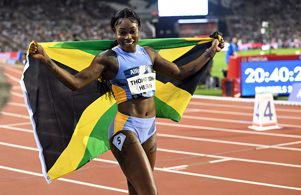 Elaine Thompson-Herah jubilantly holds the Jamaican flag after a race, wearing her athletic gear with bib number 5