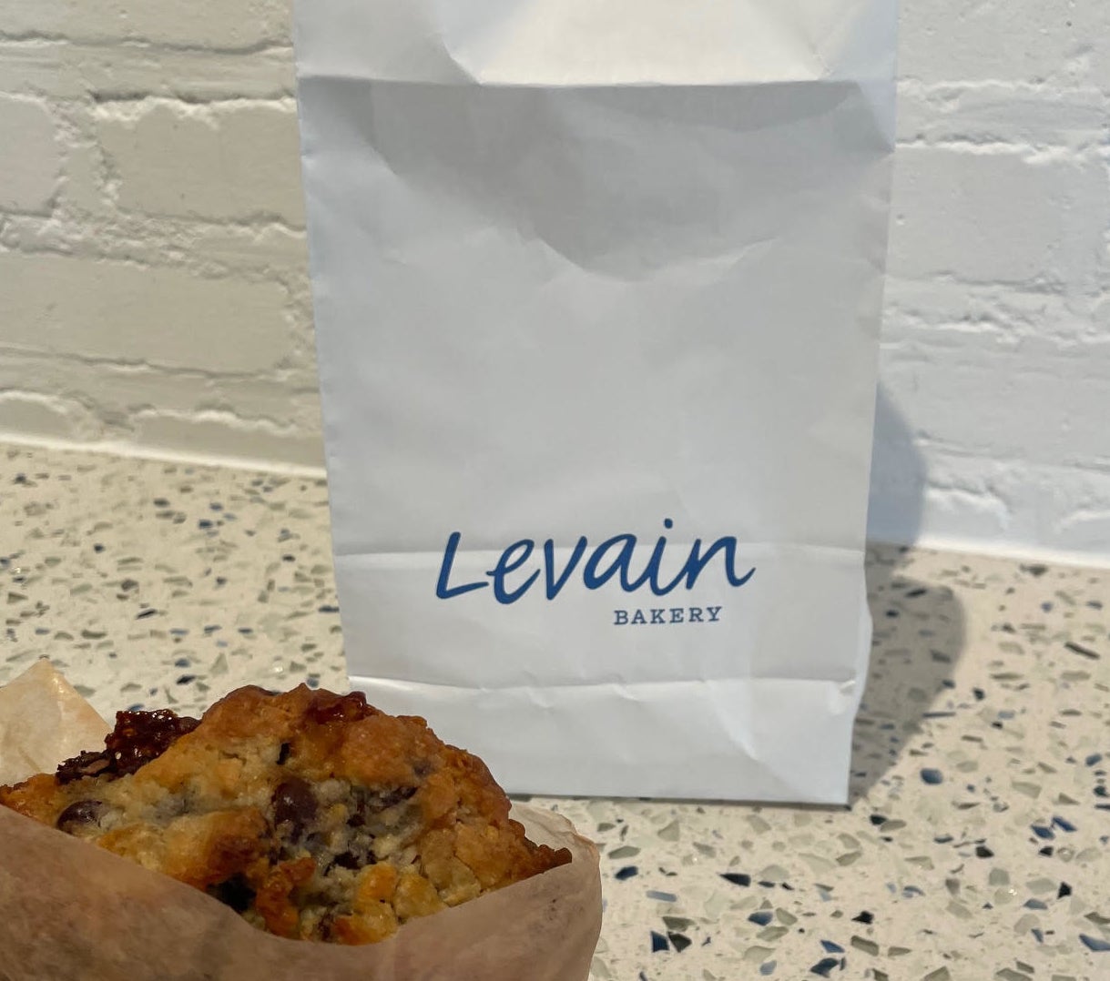 A person&#x27;s hand holding a chocolate chip cookie in front of a Levain Bakery bag