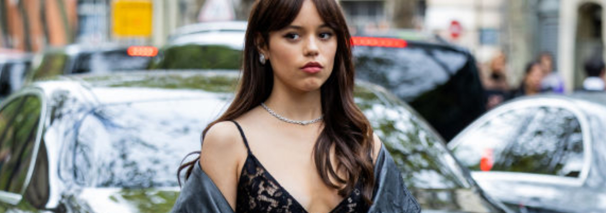 Jenna Ortega in a lace top and leather jacket, standing in front of parked cars