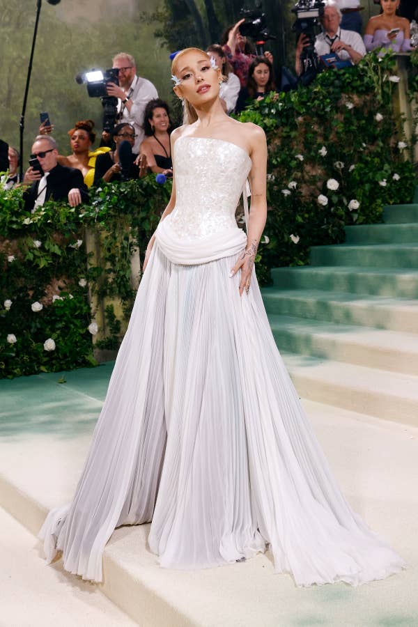 Ariana Grande in elegant one-shoulder gown with pleated skirt on steps, photographers in background