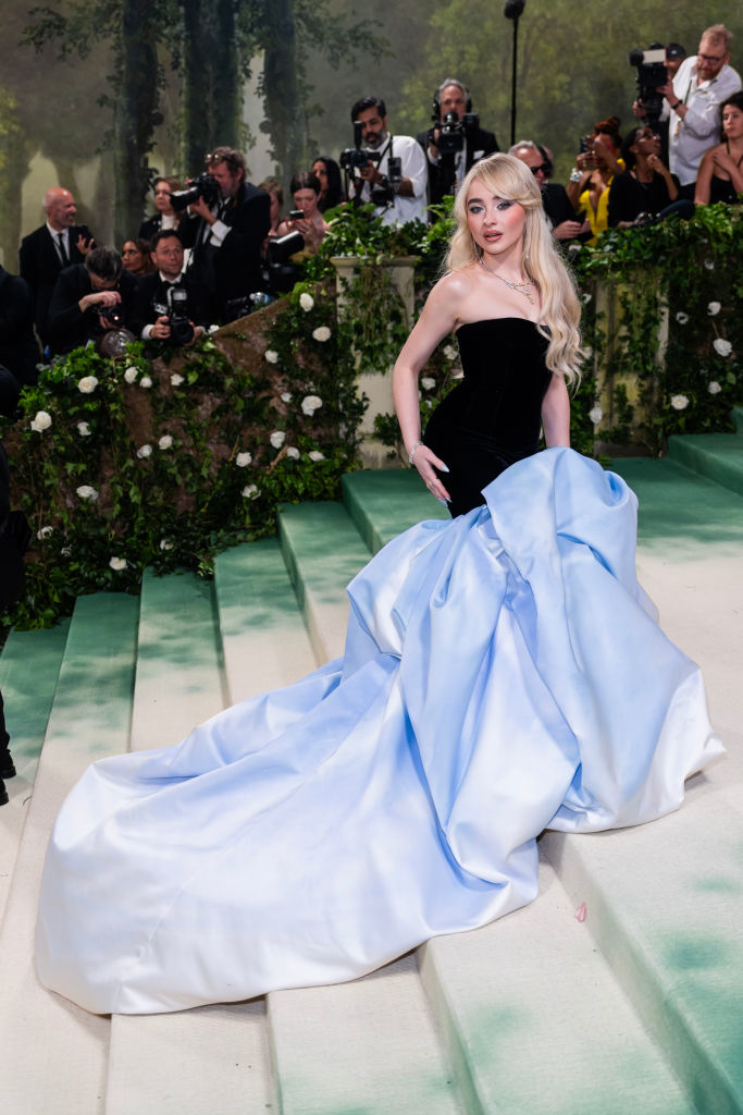 Sabrina in sleeveless top and flowing skirt on carpet, photographers in background