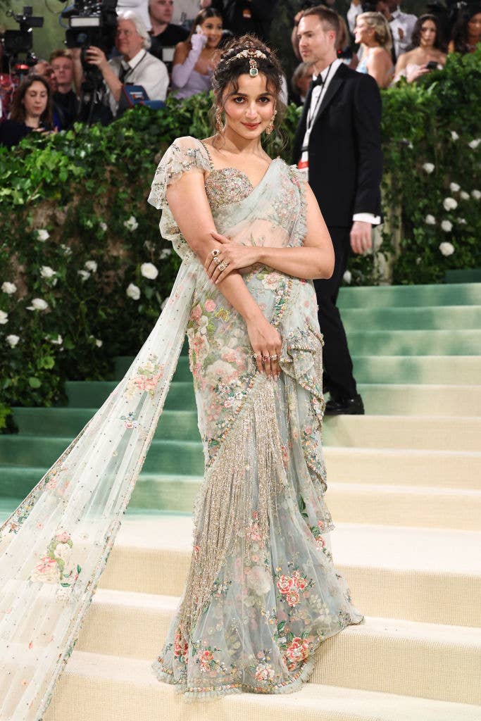 Alia on red carpet in a floral embroidered gown with a sheer cape, posing with hand on hip