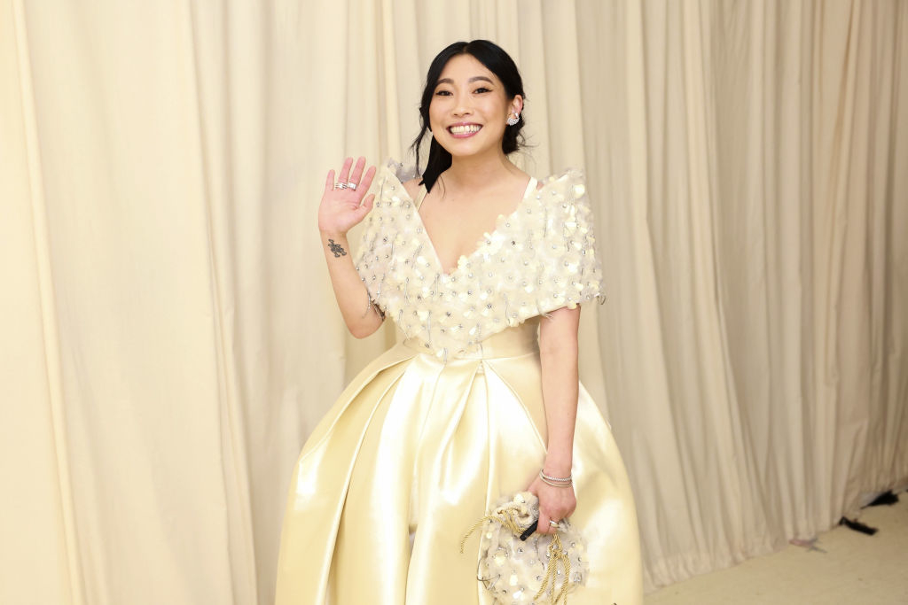 Awkwafina waving, dressed in a gown with a floral top and voluminous skirt, standing in front of a curtain