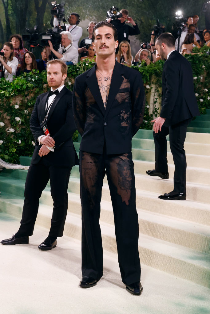Damiano in sheer, lace-patterned suit stands on stairs with attendee behind him
