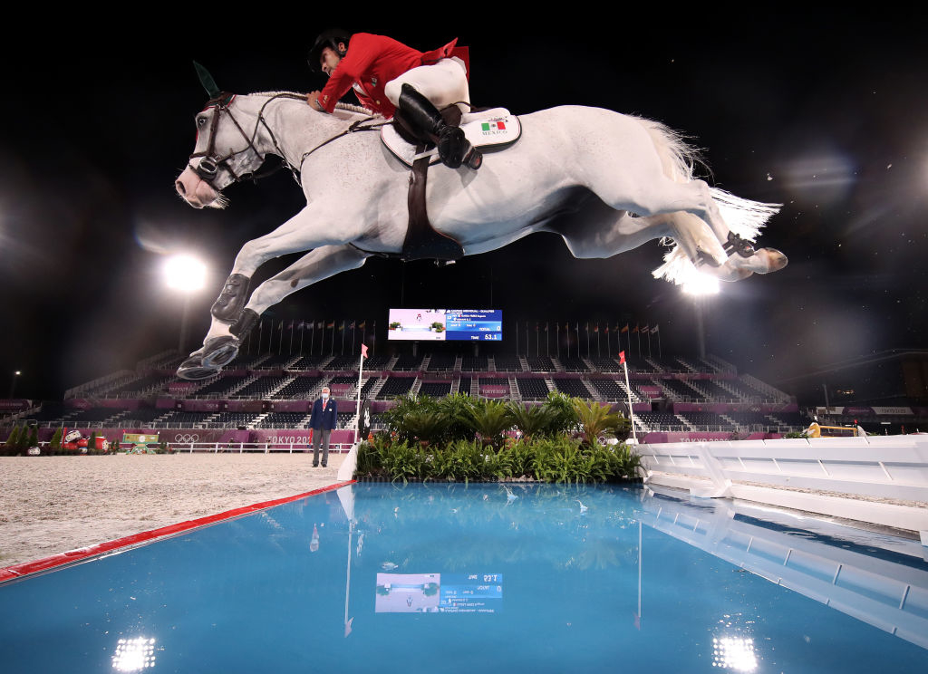 Equestrian jumping a hurdle with a grey horse at a night event, officials observing in the background