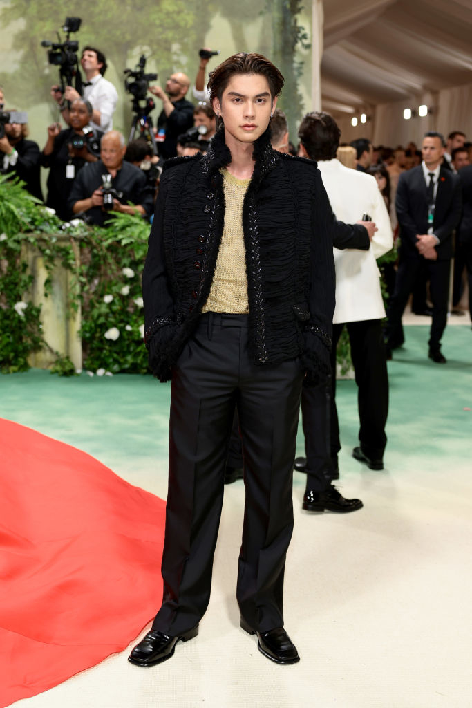 Bright at an event wearing a textured black jacket, gold top, and black trousers, with photographers in the background