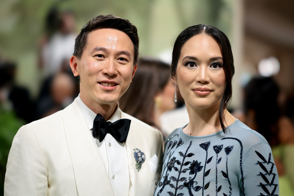 Shou and Vivan posing, one in a white tuxedo with a floral pin, the other in a sheer dress with a floral pattern