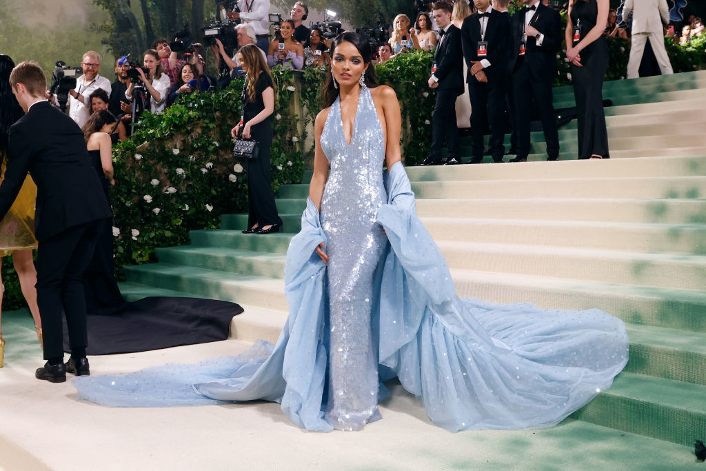 Rachel on red carpet in a glittering gown with a long train, posing on stairs with onlookers and photographers in the background