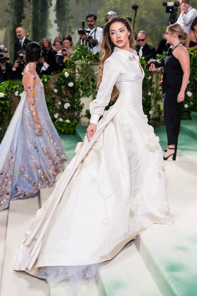 Madelyn in an elaborate dress with floral accents, long sleeves, long train, and collar posing on a carpet, photographers in the background