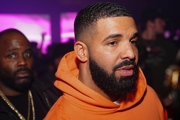 Drake in an orange hoodie at an event, with another person visible in the background