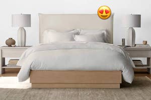 Elegantly made bed with light wood base and plush headboard, two nightstands with lamps and decor, heart eyes emoji above the bed