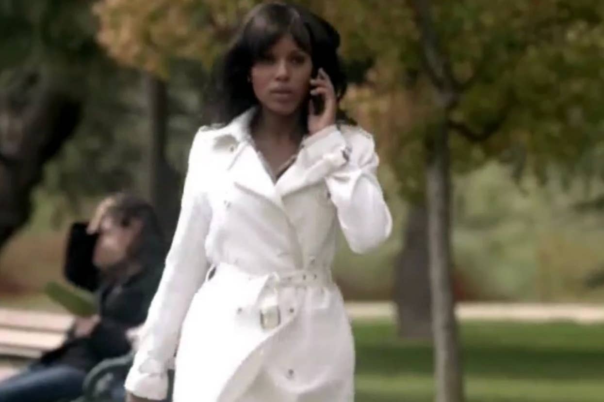 Woman in a white trench coat on a phone call, walking in a park with trees and a sitting person in the background