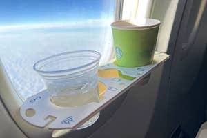 A cup of Starbucks coffee and a plastic cup on a fold-down tray attached to airplane window