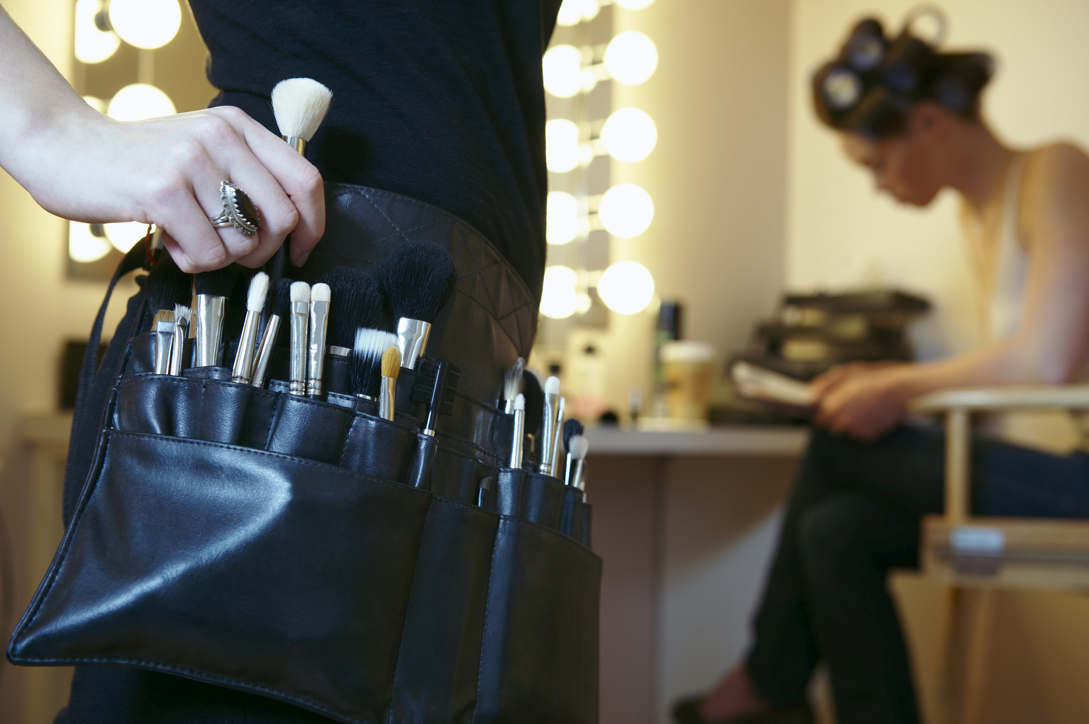 Makeup artist selecting a brush from belt pouch, client in background, interior setting
