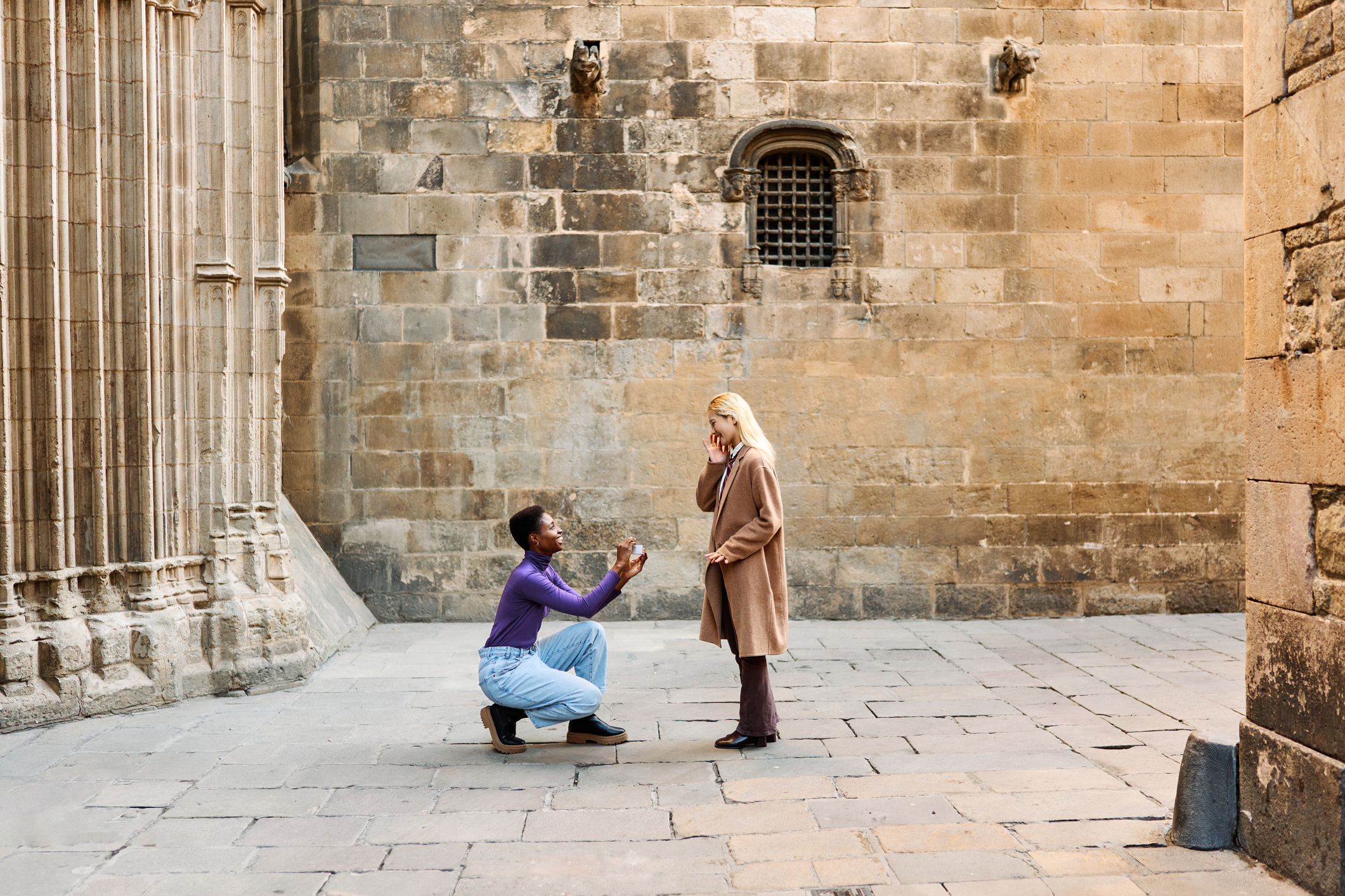 Man on one knee proposing to a woman in a public square. Both are smiling