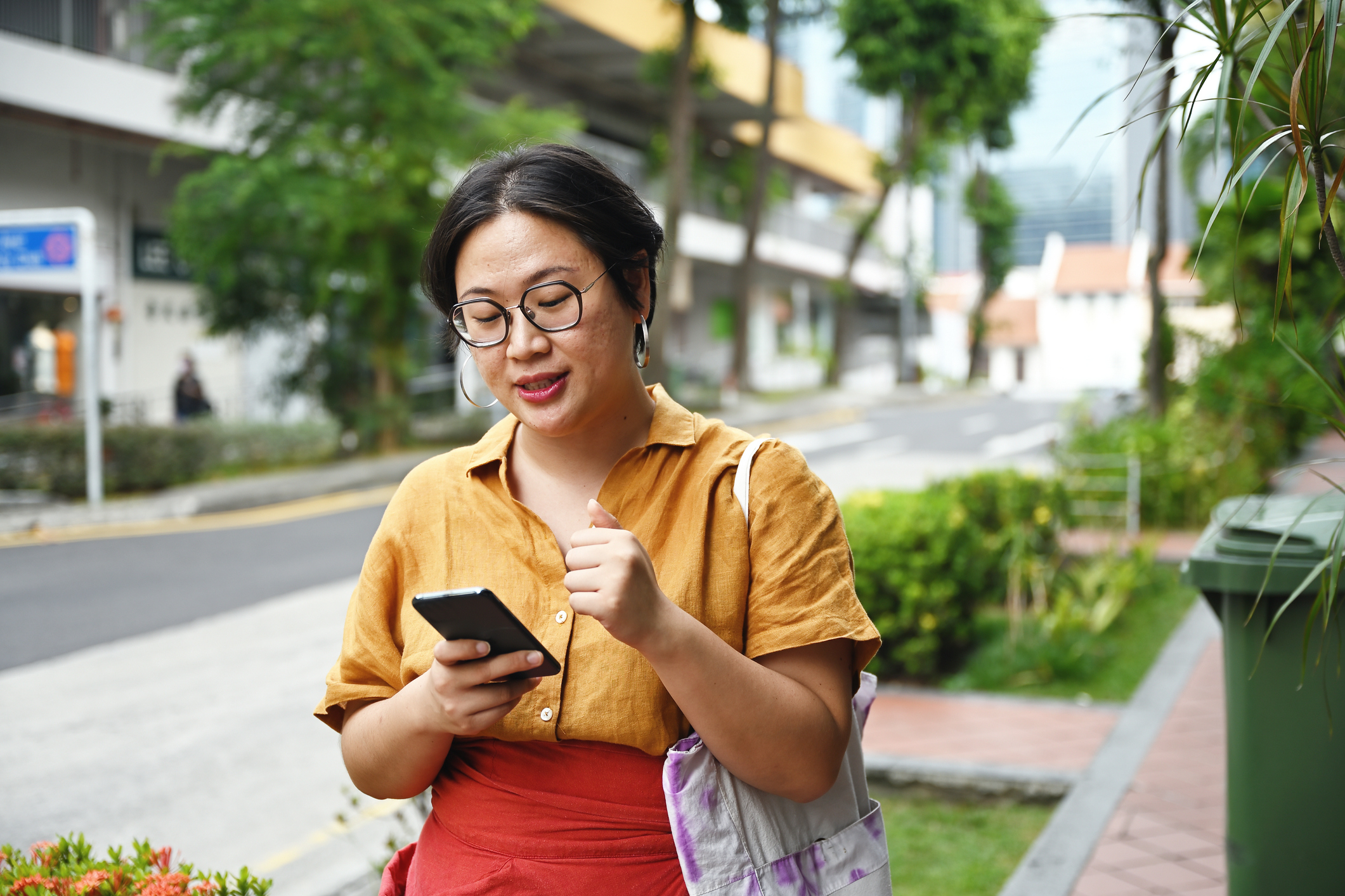 Woman in glasses using phone, carrying a bag, standing on a sidewalk