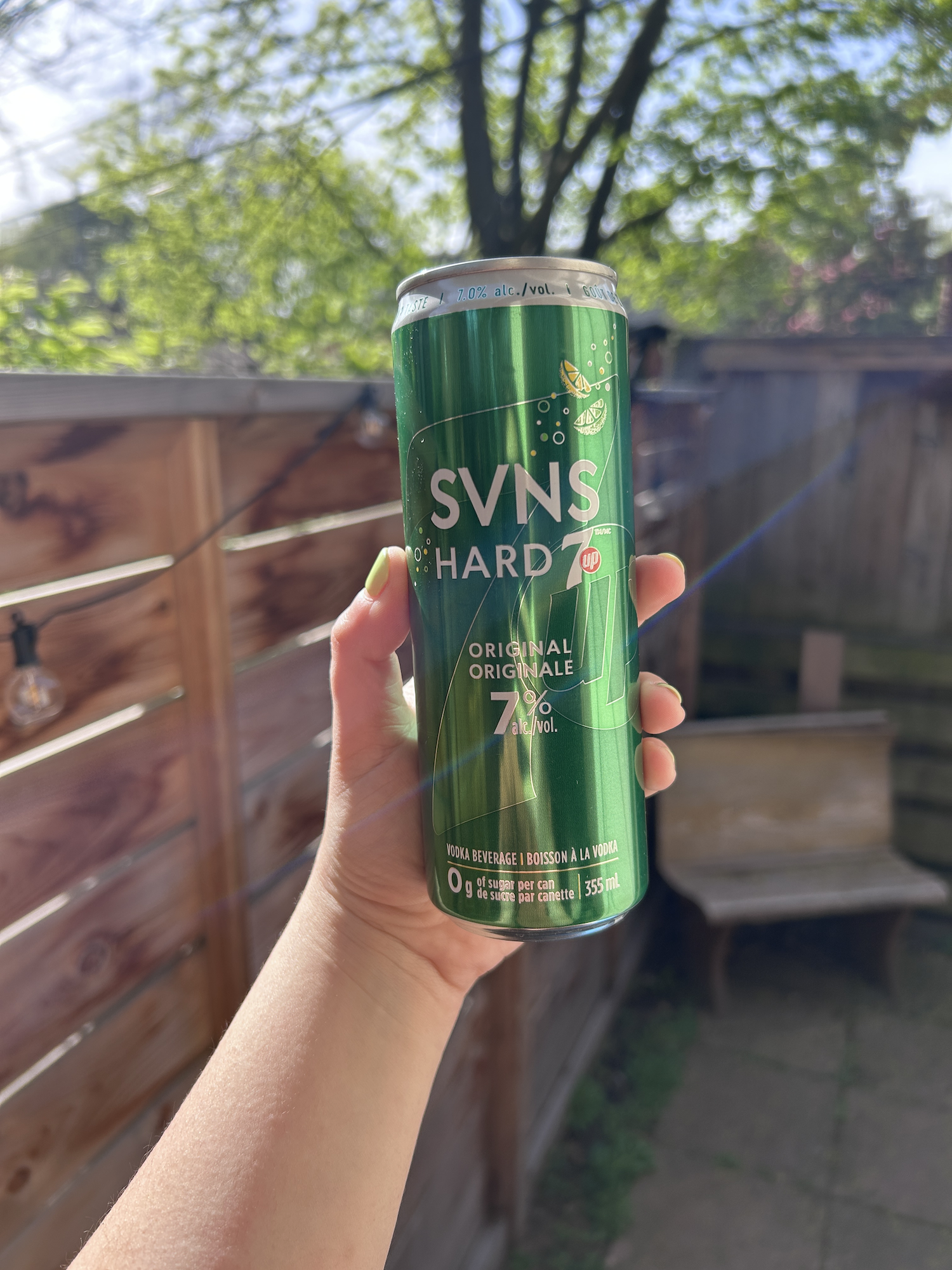 Person holding a can of SVNS HARD Seltzer outdoors with a wooden fence and foliage in the background