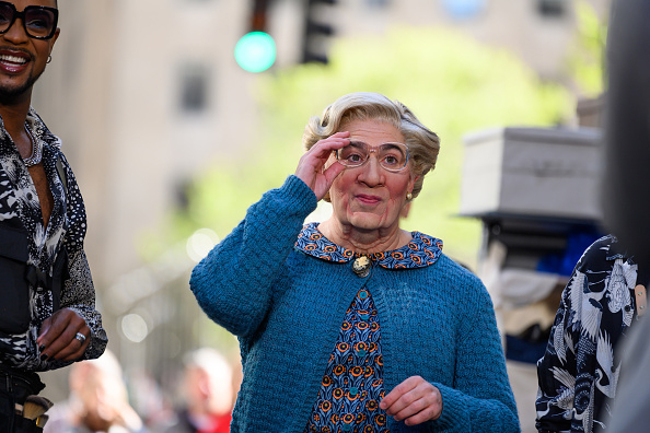 Elderly woman with glasses adjusts her eyewear, beaming, with a bystander in a patterned jacket to the left