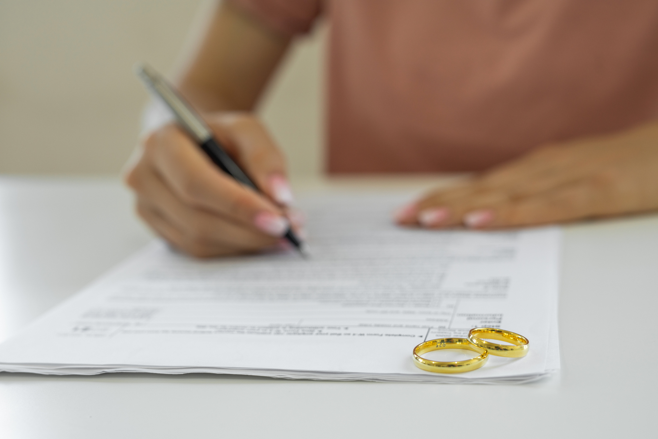 Person signing document beside two wedding rings, implying a theme of marriage commitment or legal aspects of relationships