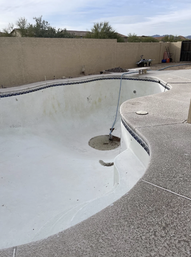 Empty in-ground swimming pool with debris and a pool skimmer, surrounded by a patio under a clear sky