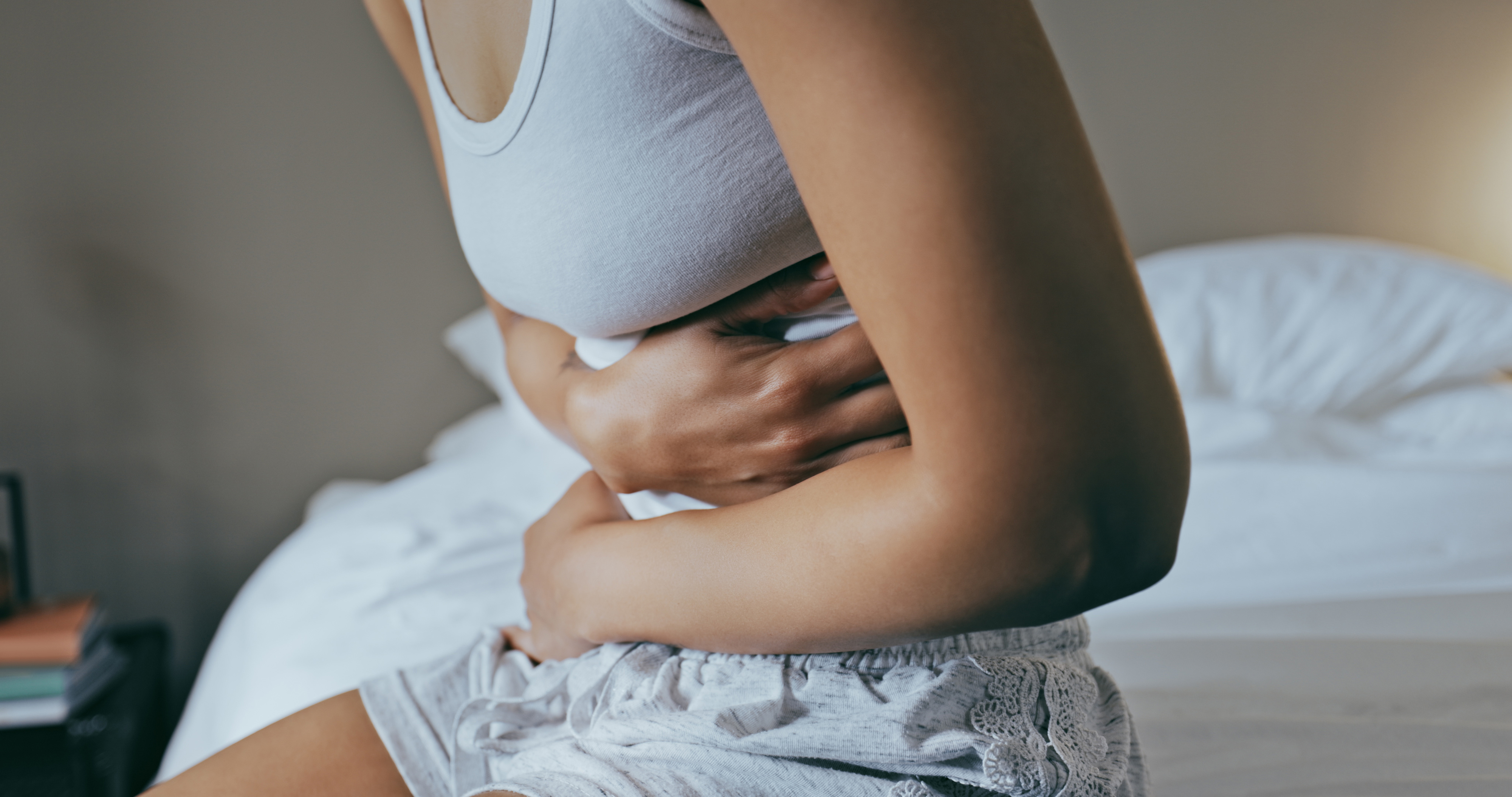 Woman experiencing stomach pain, sitting on bed, holding abdomen, with a wellness device on back