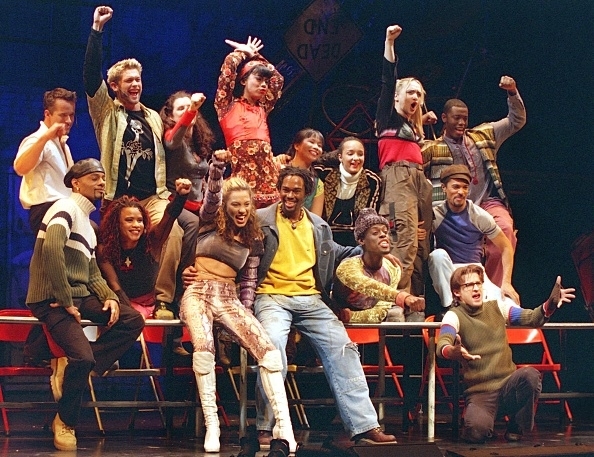 Cast of a stage musical mid-performance, expressing joy with raised arms and enthusiastic poses