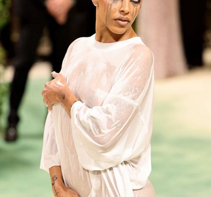 Doja Cat at an event wearing a sheer, draped outfit and sporting short blonde hair with artistic makeup