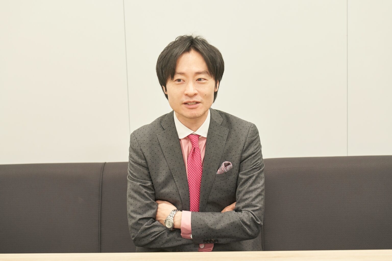 スーツとピンクのネクタイを着た男性がソファに腕を組んで座っている。