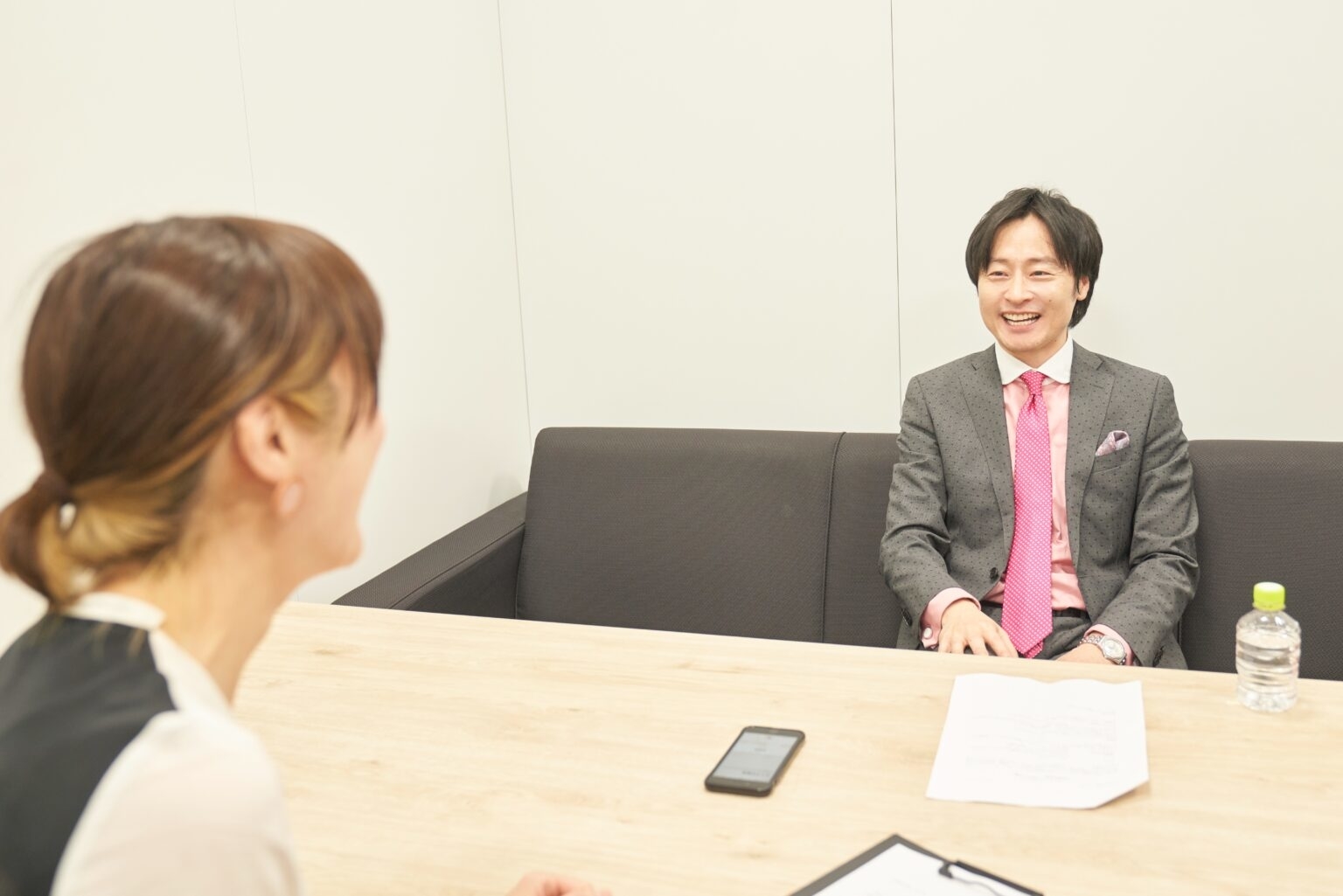 二人がオフィスで対話している。男性がスーツを着て笑顔、女性が背中側から見える。