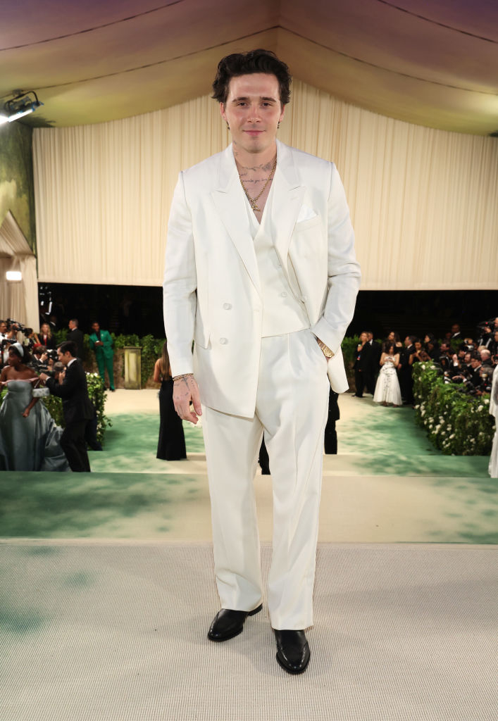 Brooklyn Beckham in a white suit with double-breasted jacket and black shoes, standing on a carpeted area with onlookers in background