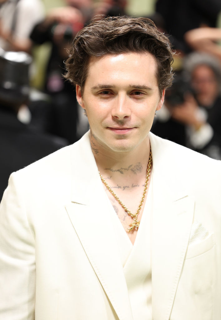 Brooklyn Beckham in a white suit with chest tattoos visible, smiling at an event