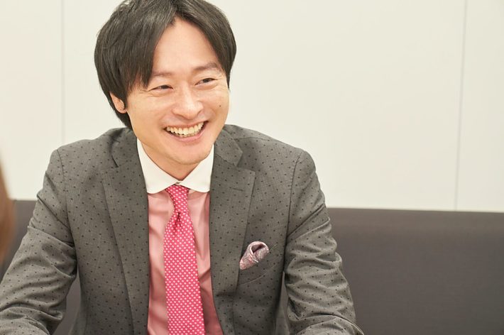 笑顔の男性がドット柄のスーツとピンクのネクタイを着用してソファに座っている。