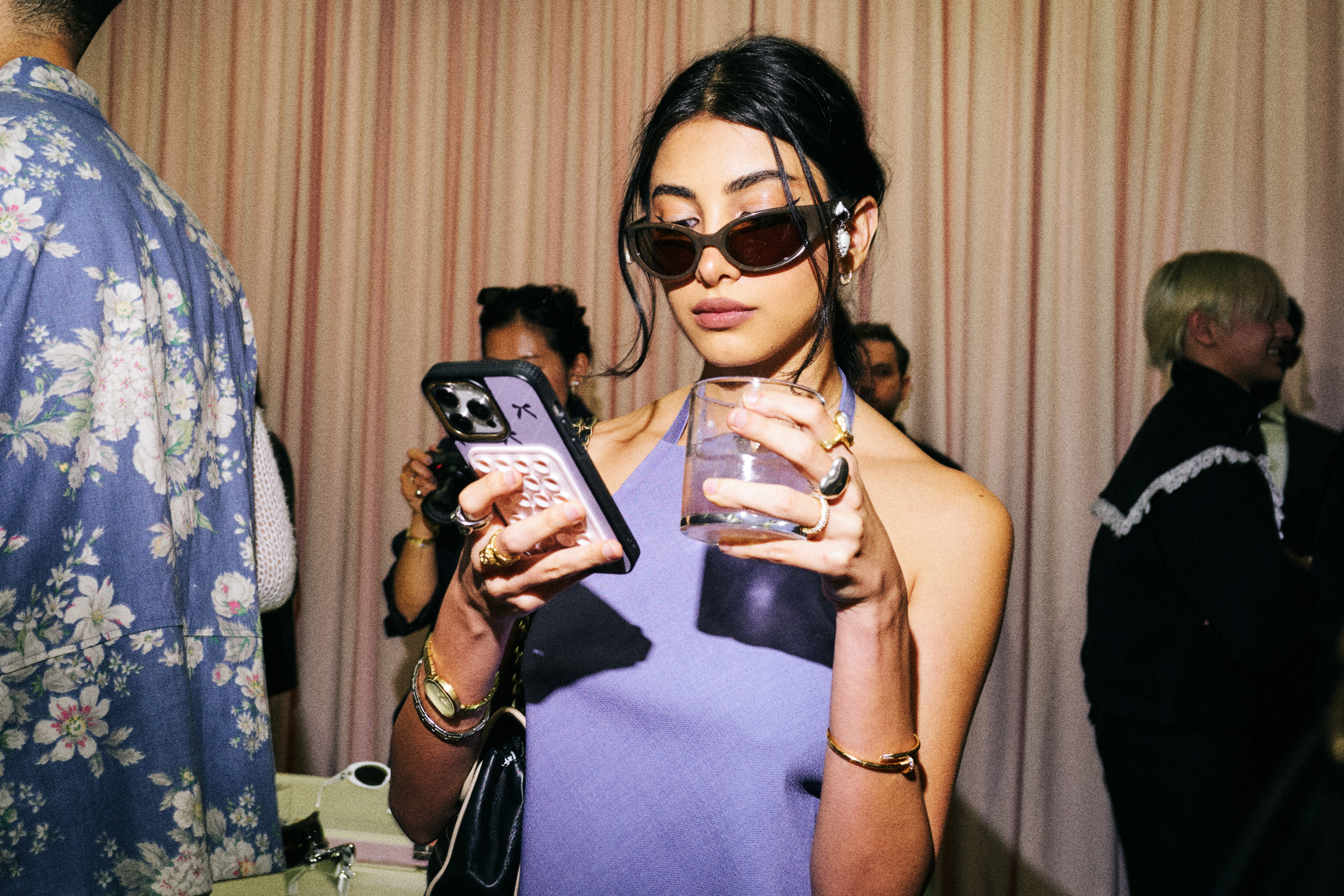 Person in a sleeveless top with sunglasses holding a phone and a drink at a social event