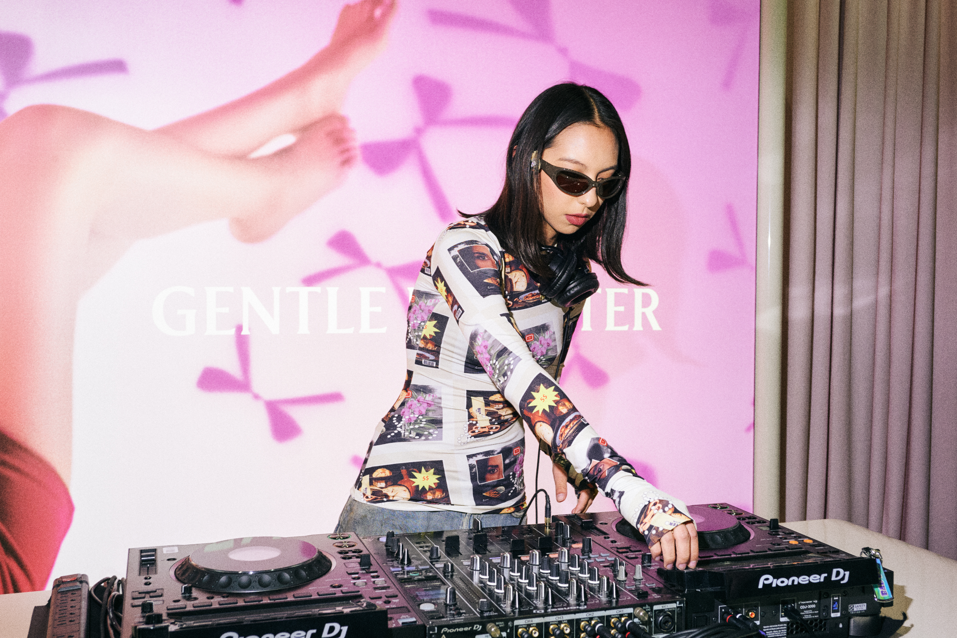 DJ wearing a printed long-sleeve top and sunglasses at a mixer beside a Gentle Monster ad