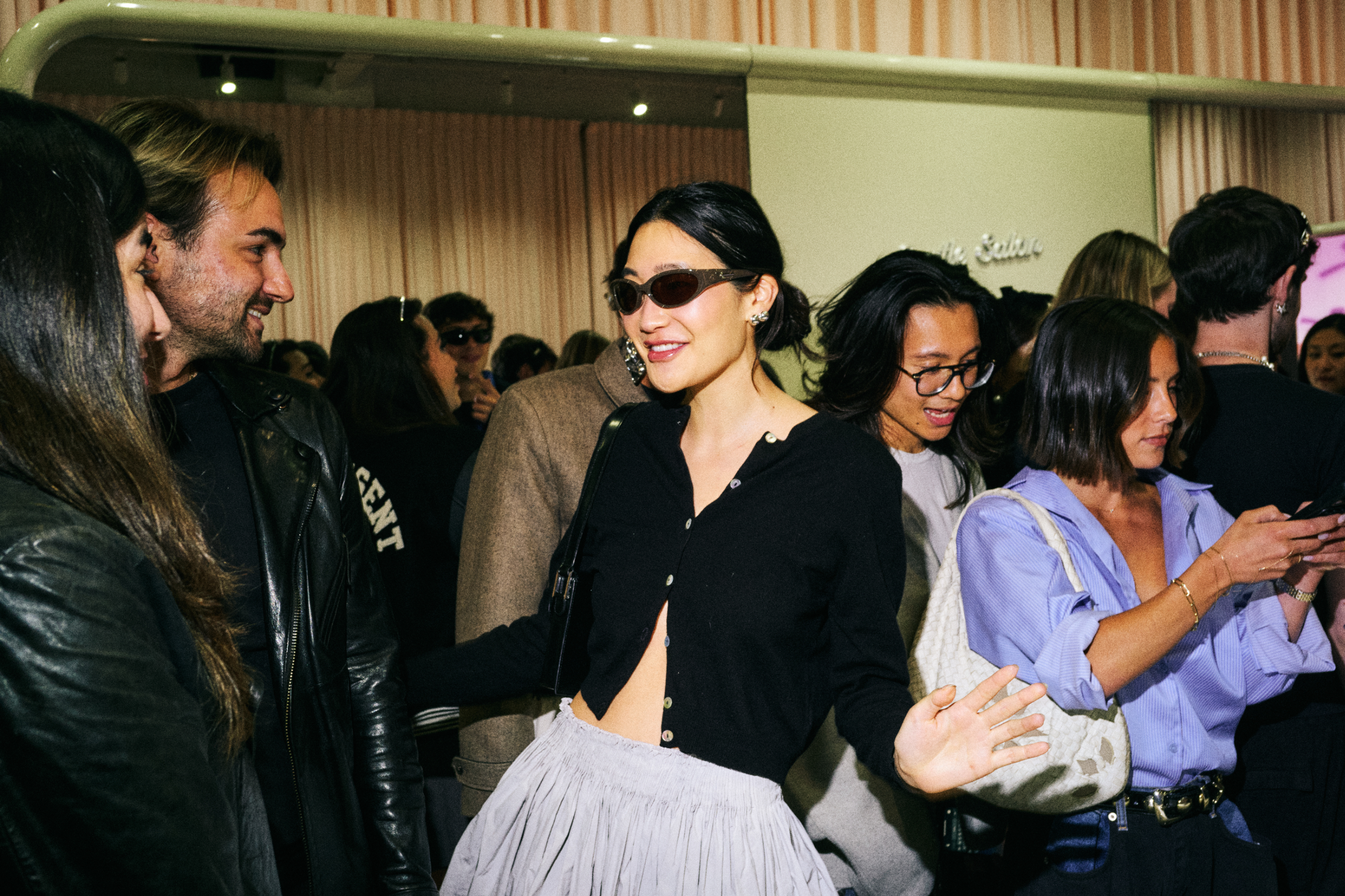 Woman in a chic black top and pleated skirt, sunglasses indoors, engaging with event attendees