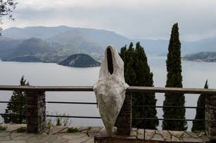 Sculpture of a figure with no head or arms overlooking a lake with mountains and cloudy sky