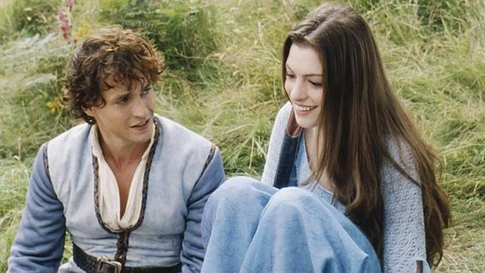 Dos personajes de una película sentados sobre la hierba, él viste camisa y chaleco, ella lleva un chal azul